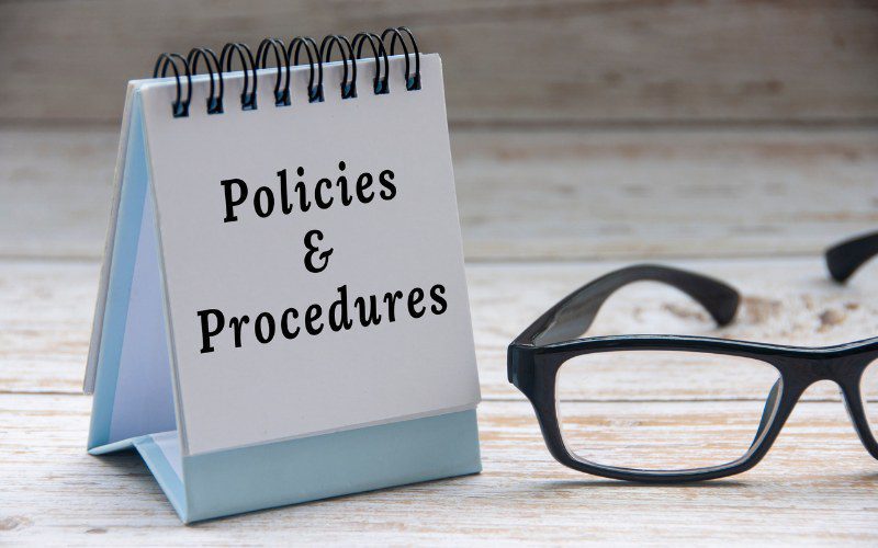 A policies and procedures sign sits on a work desk next to a pair of reading glasses.